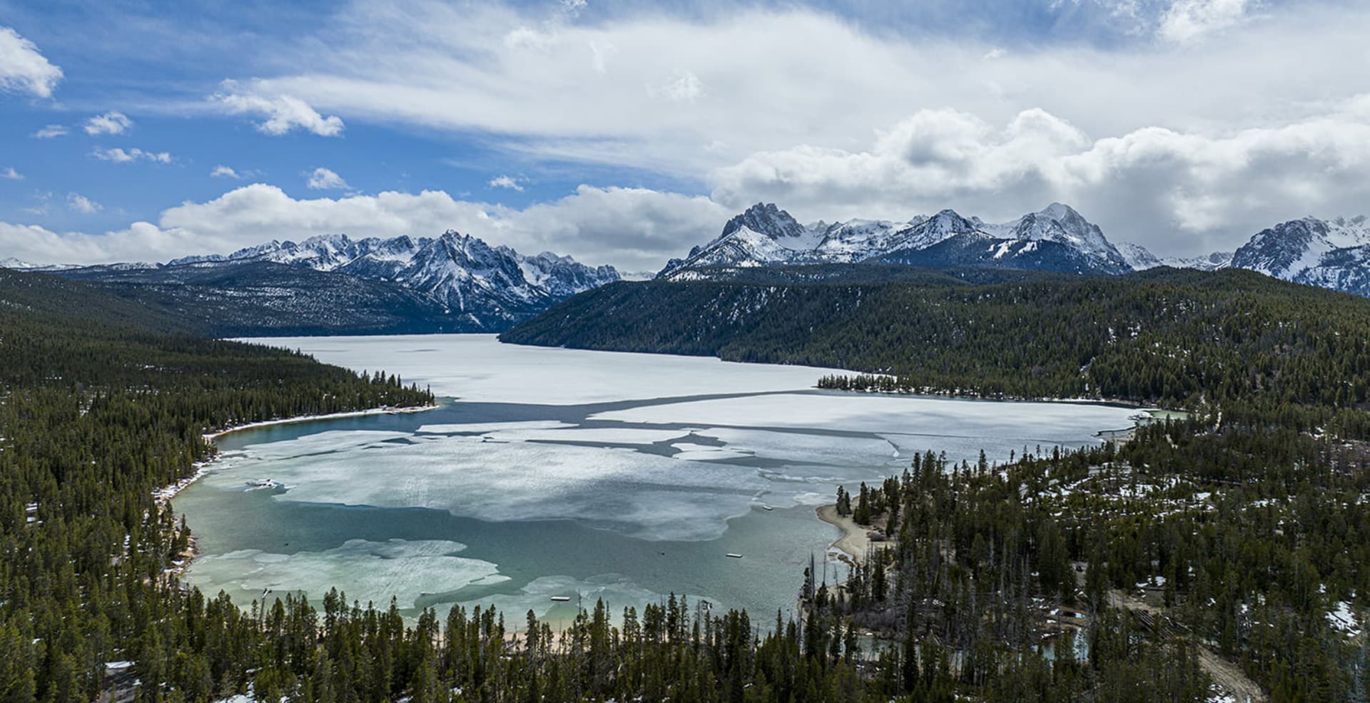 Aerial view of a partially frozen lake surrounded by snow-capped mountains and dense forests under a cloudy sky.